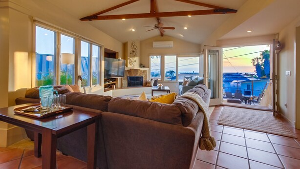 A spacious living area with a large couch, a dining table, and a television. There is a balcony with a view of the sky and palm trees. - Experience coastal charm near Moonlight State Beach (Encinitas)
