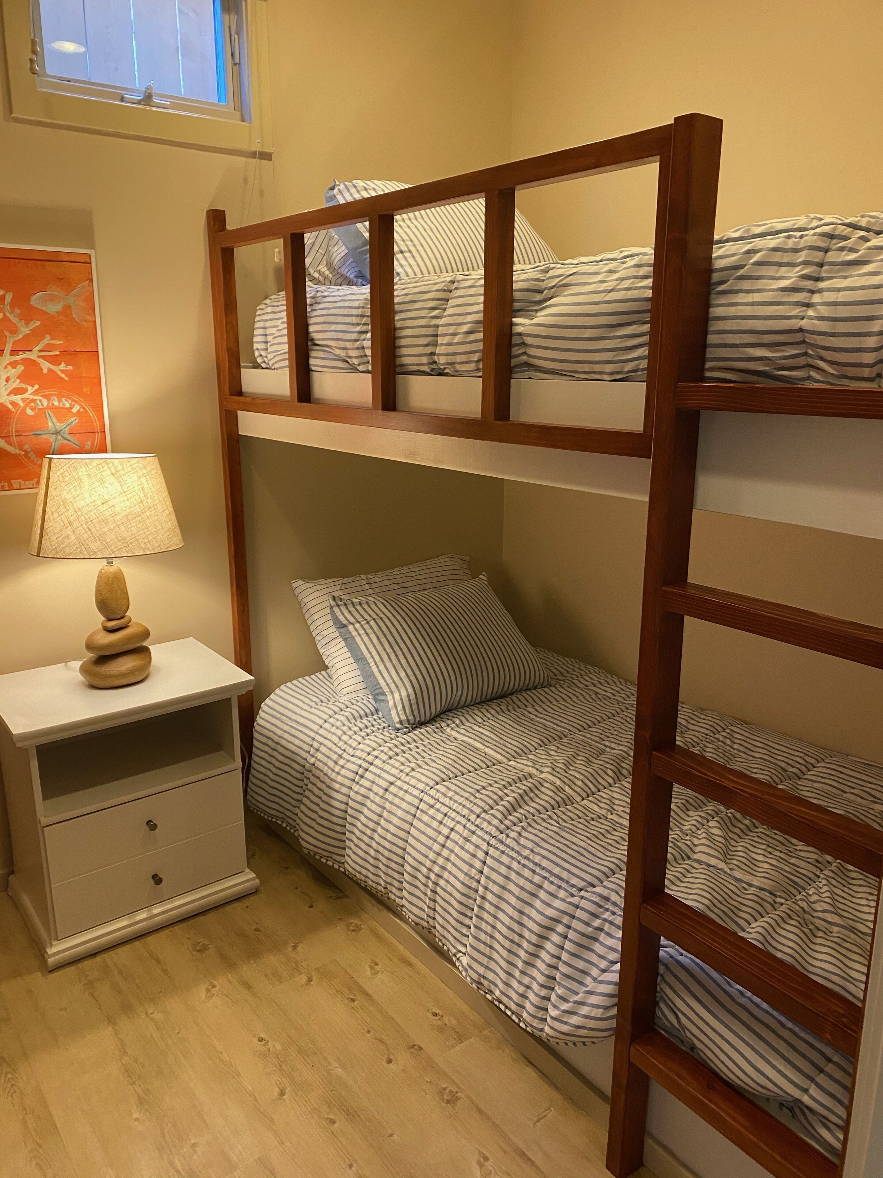 Une chambre avec un lit superposé, du linge de lit à rayures, une échelle en bois, une table de chevet avec une lampe et un tableau encadré au mur.