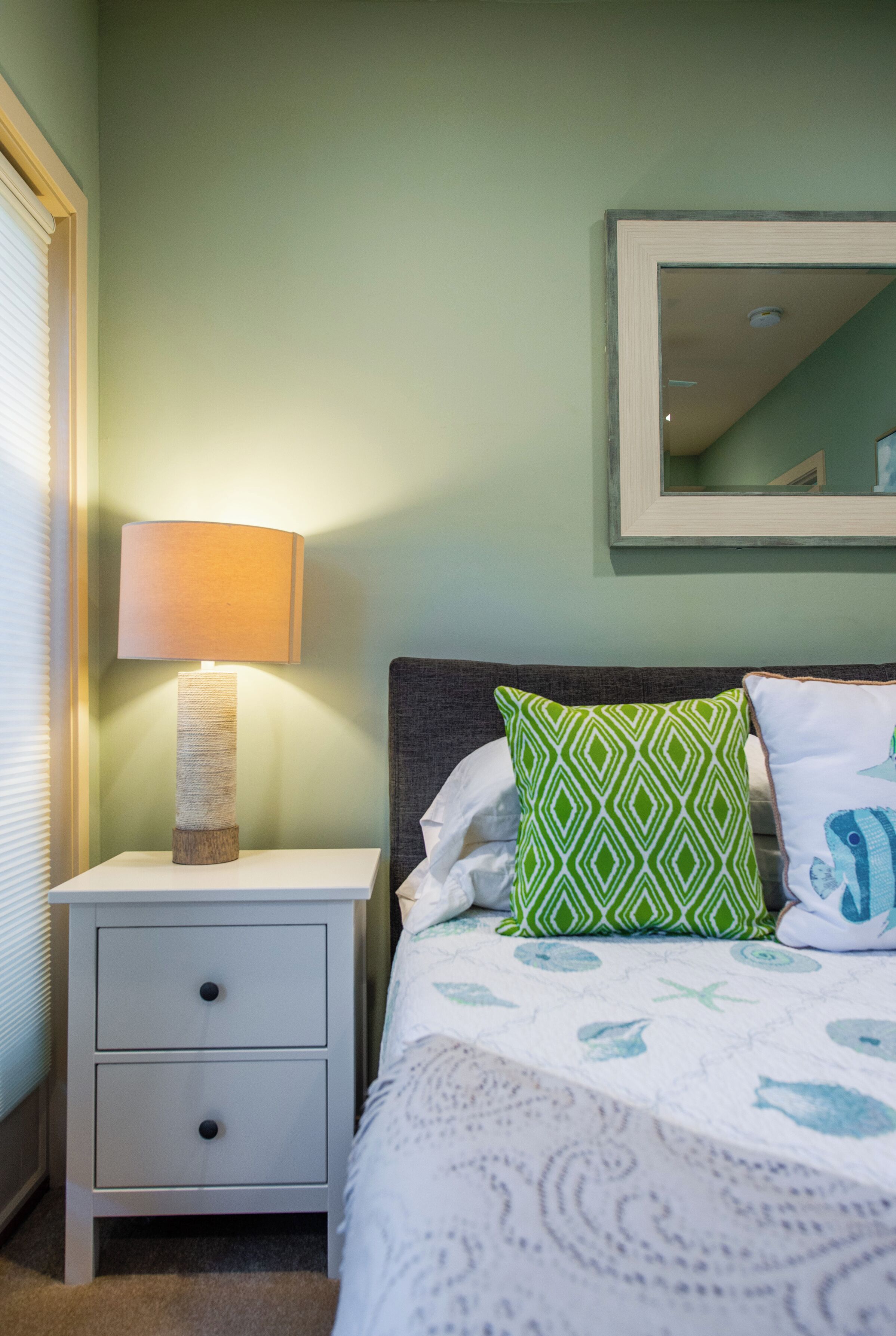 Una cama bien hecha con una colcha estampada, una lámpara de noche, un almohadón verde con diseño y un espejo en la pared.
