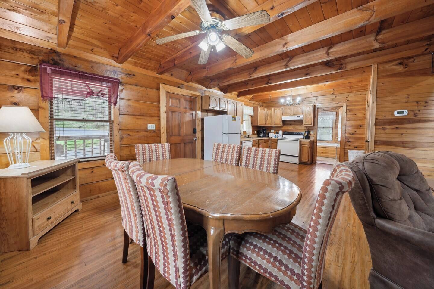 A wooden cabin interior with a dining table, chairs, a sofa, a ceiling fan, and a kitchen area.