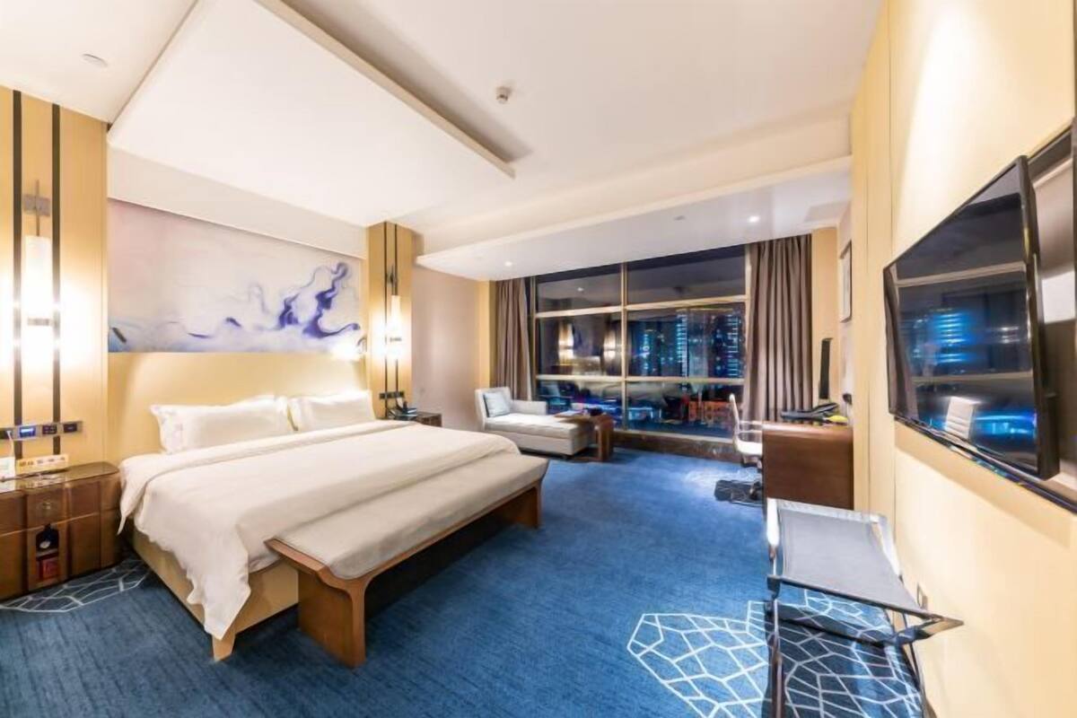 A hotel room with a large bed, a desk, a chair, a sofa, a large window with a city view, and a painting on the wall.