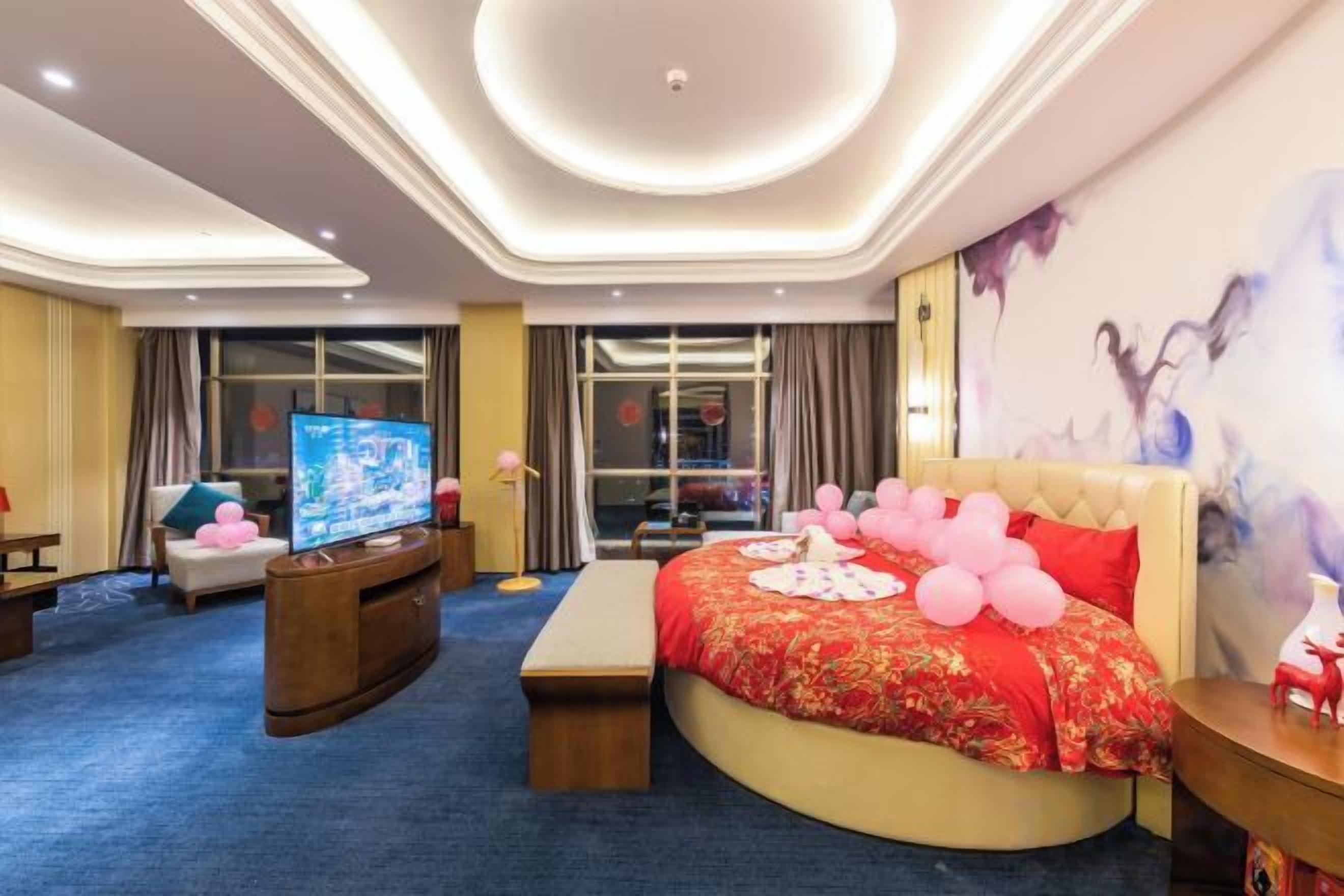 A hotel room with a large bed, a television, and a view of the cityscape through the windows.