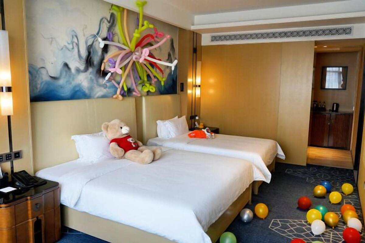 A hotel room with two beds, a large abstract painting, a teddy bear, and colorful balloons on the floor.