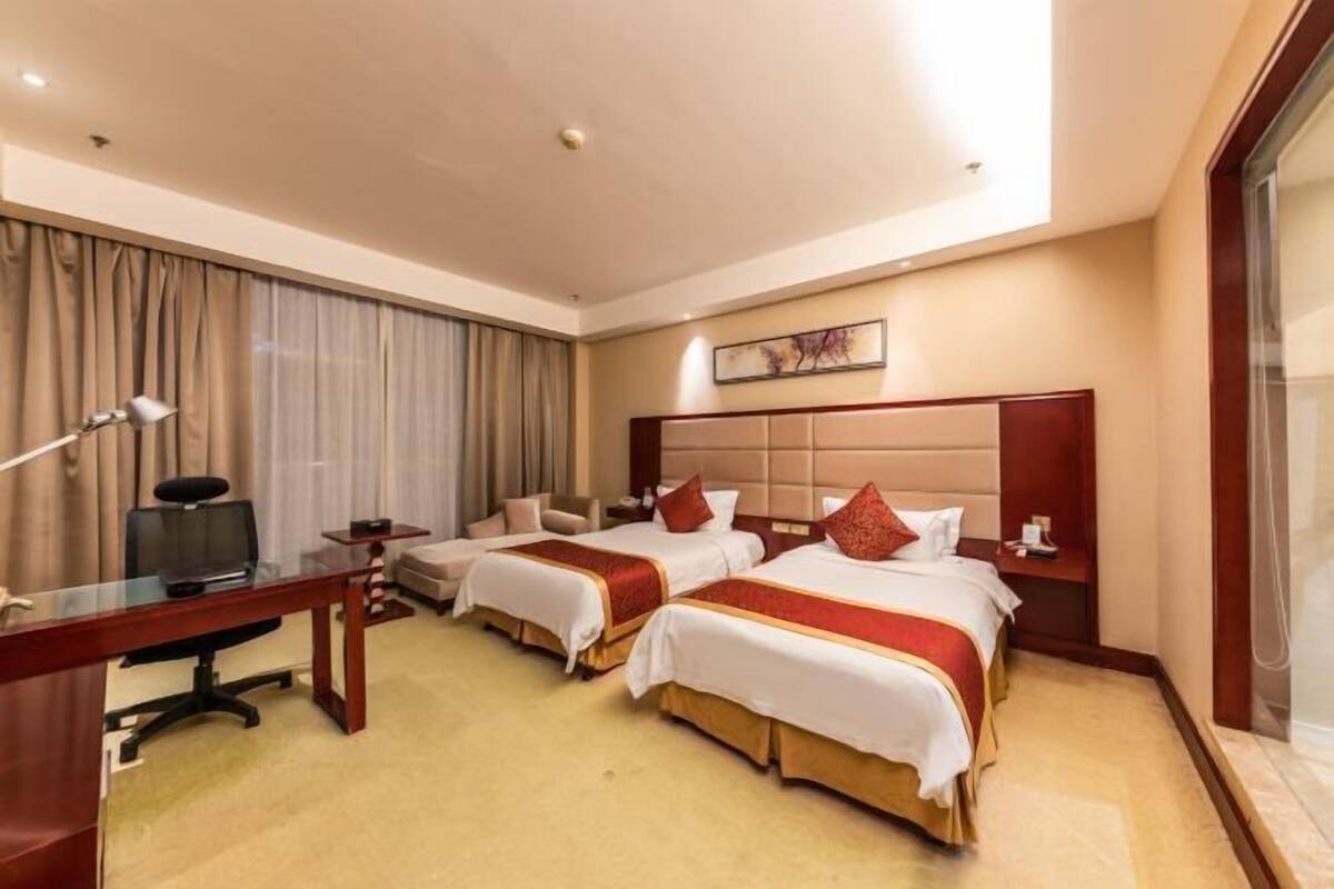 A hotel room with two beds, a desk, a chair, a television, and a large window with curtains.