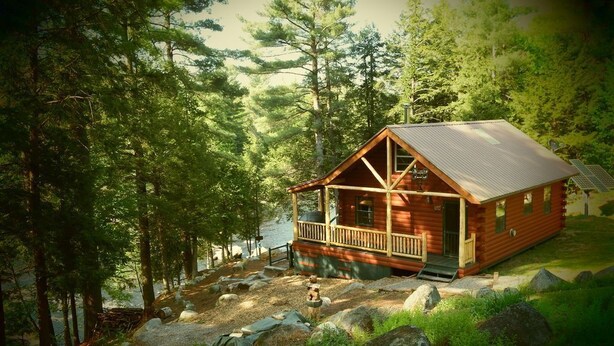 A cabin with a porch, surrounded by trees and rocks. - Adirondack/Oswegatchie Riverfront Cabin (Harrisville)