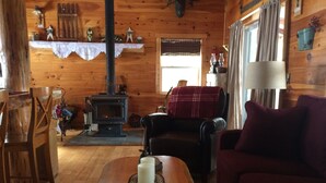 A cozy cabin interior with a wooden floor, a sofa, a chair, a table with candles, a fireplace, and a window with curtains. - Adirondack/Oswegatchie Riverfront Cabin (Harrisville)