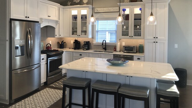 Una cocina moderna con armarios blancos, electrodomésticos de acero inoxidable, una isla grande con taburetes de bar, iluminación colgante y un ventanal con persianas.