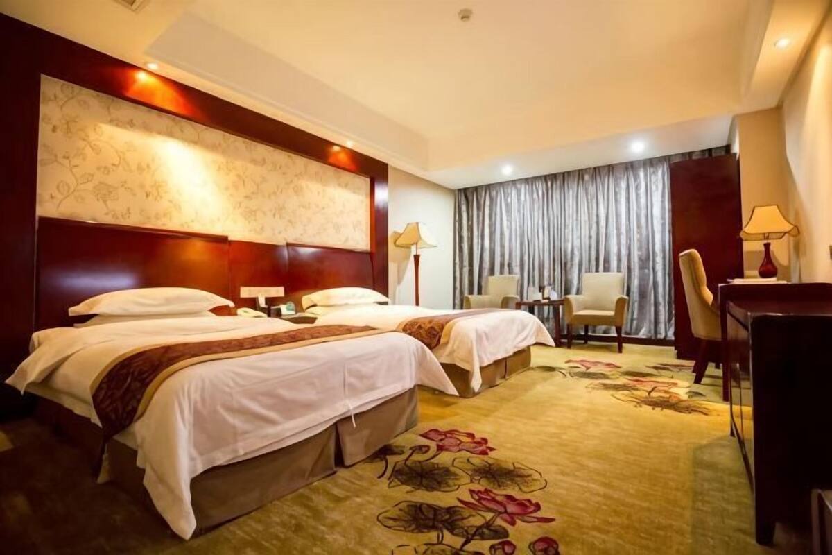 Habitación de hotel con dos camas, un escritorio, una silla, una lámpara y un ventanal grande con cortinas.