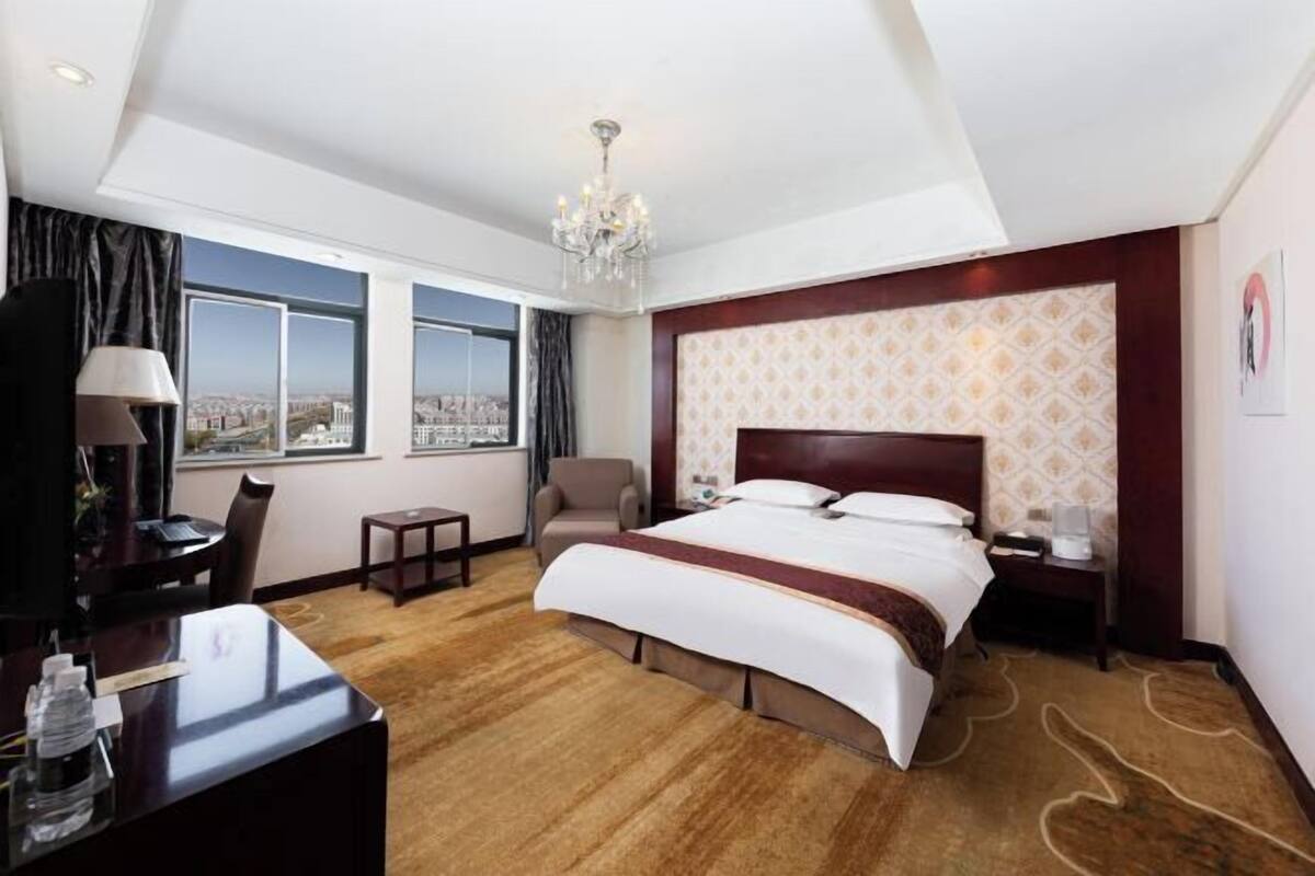 Habitación de hotel con una cama grande, un escritorio, una silla, una mesita pequeña, una mesita de noche, una lámpara, un candelabro y ventanas amplias con vistas a la ciudad.