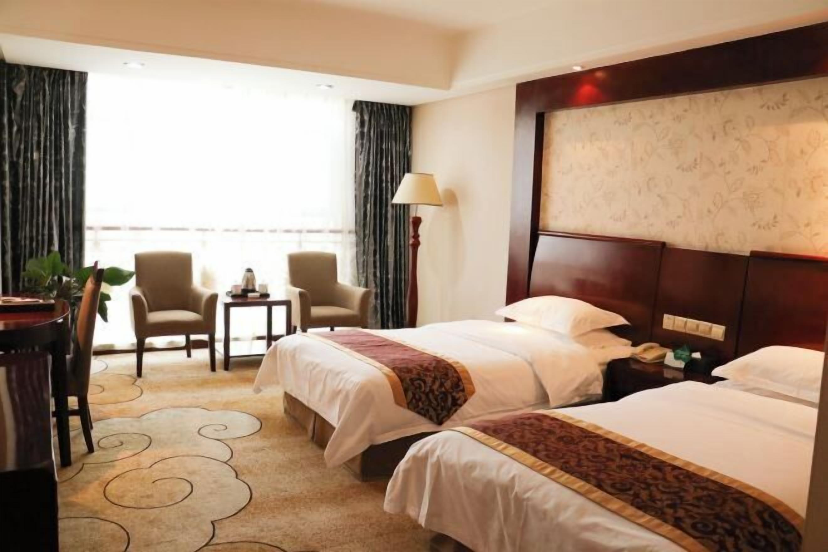 A hotel room with two beds, a desk, chairs, a lamp, and a view of the outdoors.