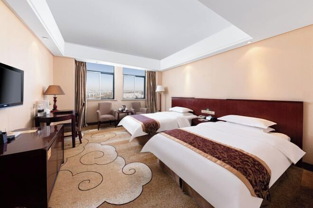 A hotel room with two beds, a desk, a chair, a TV, and a view of the city.