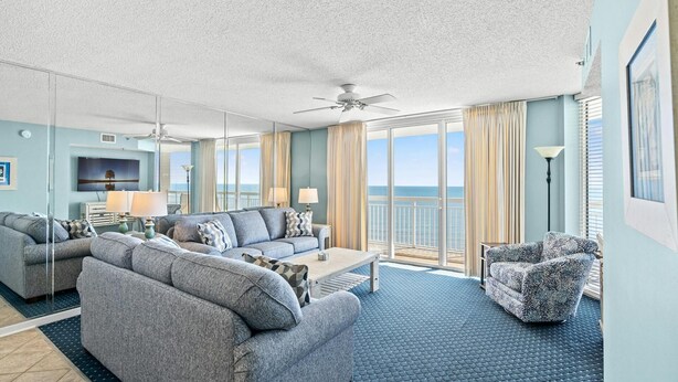 Ein geräumiges Wohnzimmer mit Sofa, Ohrensessel und Couchtisch, einem Deckenventilator und einem großen Fenster mit Meerblick.