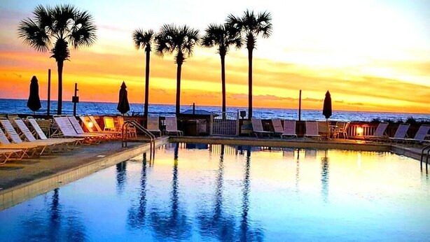 A poolside area with lounge chairs and palm trees, overlooking the ocean at sunset.