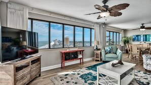 A spacious living room with a large TV, wooden entertainment unit, and a city view through large windows.