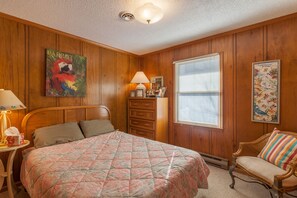 Ein Schlafzimmer mit einer holzverkleideten Wand, einem Bett mit floralem Überwurf, einem Schminktisch, einem Sessel und einem Fenster mit Jalousien.