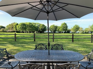 A round metal table with intricate patterns, surrounded by matching chairs, under a large umbrella, set in a grassy area with a wooden fence and trees in the background.