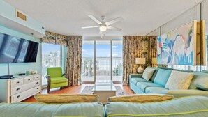 A modern living room with a flat-screen TV, a green armchair, a blue sofa, and a balcony with a view. - Upscale Oceanfront Condo, Lazy River, BBQ Grill | Crescent Shores - S 107 (North Myrtle Beach)