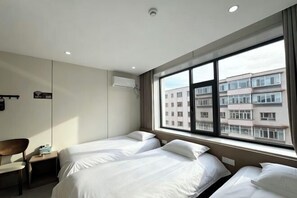 A hotel room with two beds, a desk, a chair, and a large window with a view of buildings.