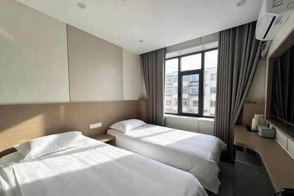 A hotel room with two beds, a window with curtains, an air conditioning unit, and a small table with a lamp.