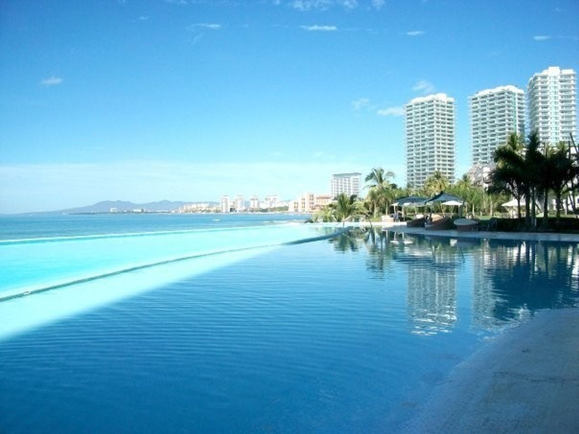 A resort pool with clear blue water, surrounded by palm trees and high-rise buildings in the background.