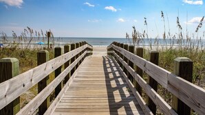 Une promenade en bois menant à une plage, avec un ciel dégagé et la mer en arrière-plan.