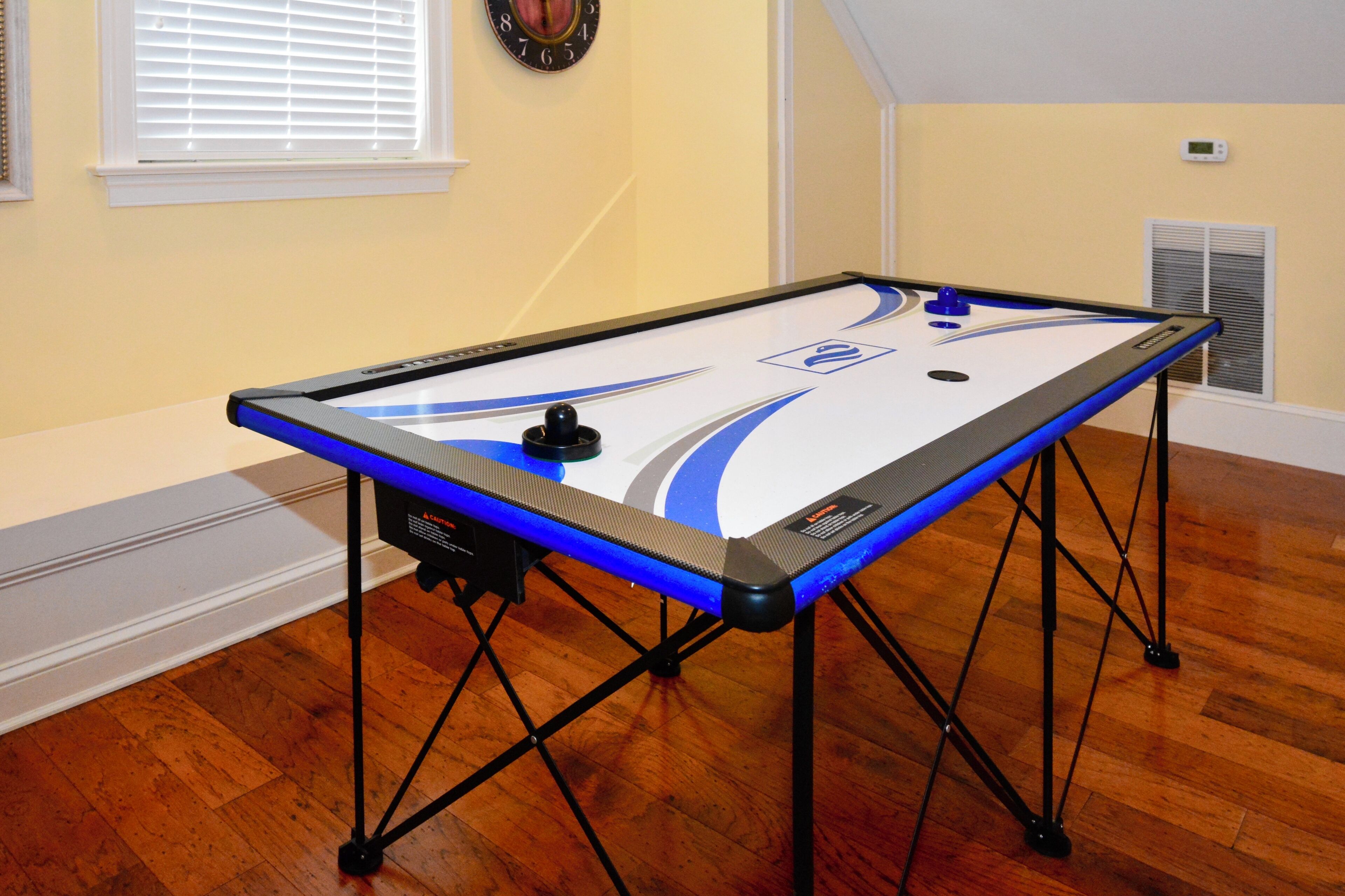 A pool table with a blue and white design, located in a room with wooden flooring and a window with blinds.