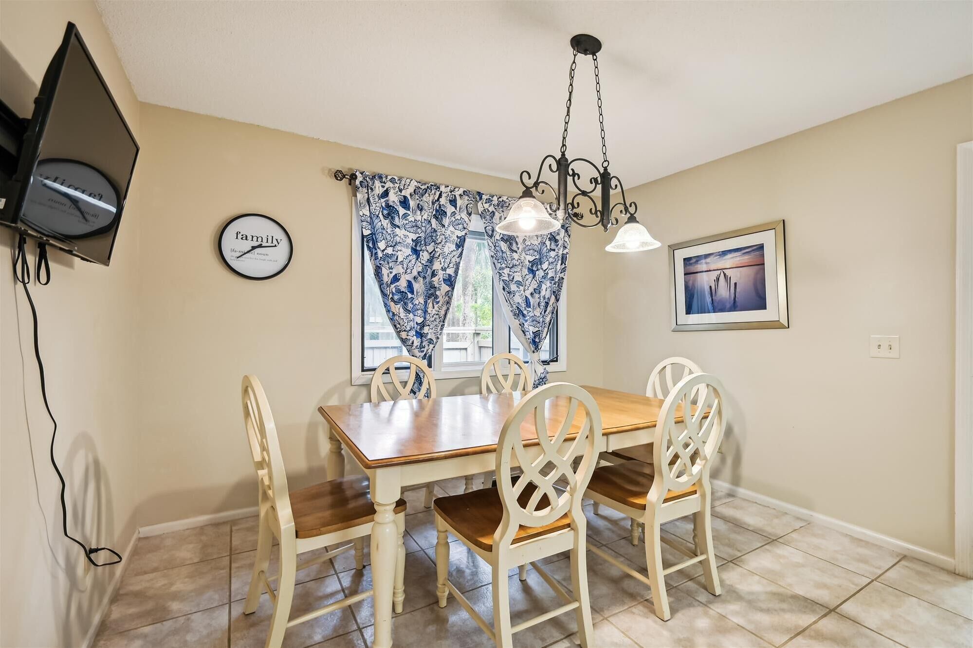 A dining area with a wooden table and white chairs, a chandelier, a clock, and a framed picture on the wall.