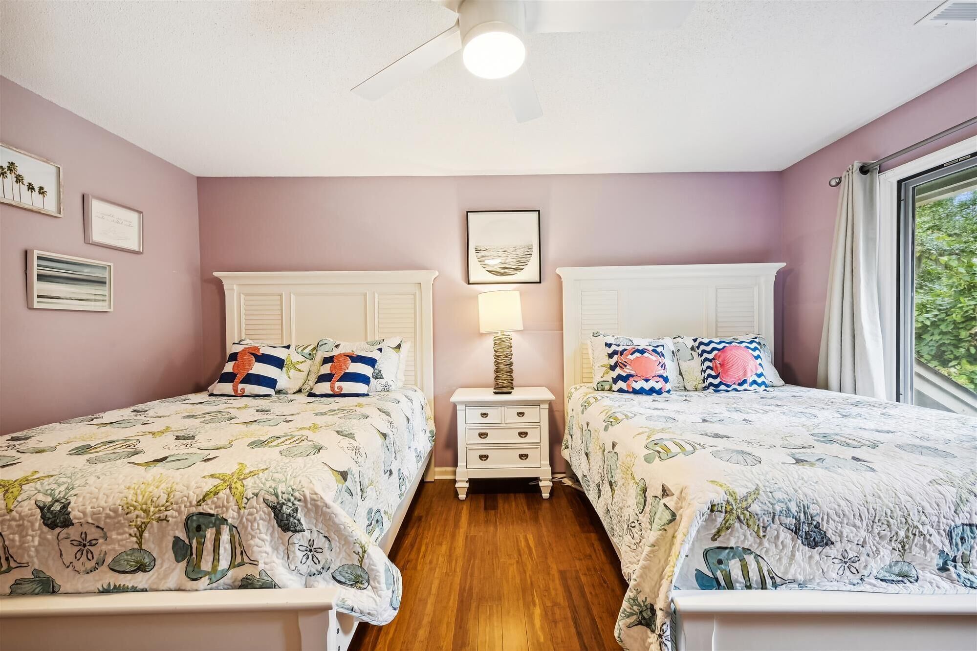 Une chambre avec deux lits, une table de chevet, un ventilateur de plafond et une fenêtre donnant sur un paysage verdoyant.