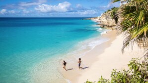A beach with clear turquoise water, a sandy shore, and a rocky cliff with vegetation. - The CLIFF - Renovated Condo in a Luxury Beachfront Resort with Spa - Restaurant (Cupecoy)