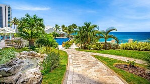A pathway leading to a pool surrounded by palm trees and greenery, with a view of the ocean and a building in the background. - The CLIFF - Renovated Condo in a Luxury Beachfront Resort with Spa - Restaurant (Cupecoy)