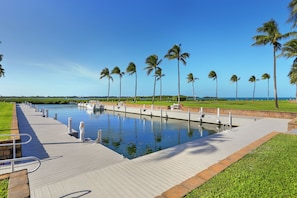 A long, narrow waterway with a wooden walkway on one side, surrounded by palm trees and a grassy area.