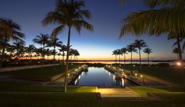 A nighttime view of a rectangular pool with a boat, surrounded by palm trees and a sunset in the background.