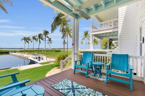A covered outdoor area with a view of a lake and palm trees, featuring a wooden deck, two blue chairs, and a small table.