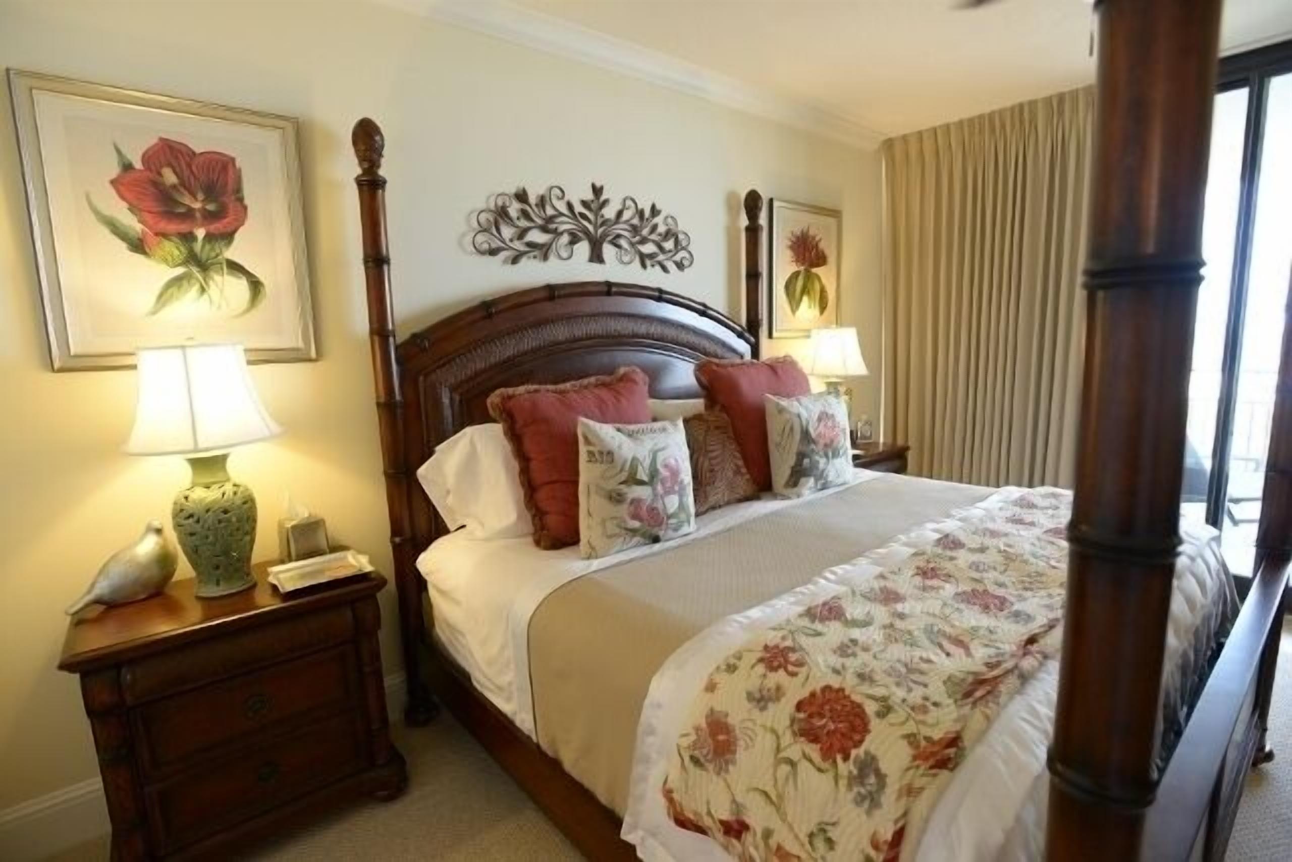 A neatly made bed with floral bedding, a wooden headboard, and decorative pillows. There is a nightstand with a lamp and a vase.