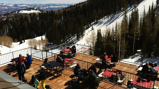 A ski resort with a wooden deck area, dining tables, and chairs, surrounded by snow-covered slopes and dense forest.