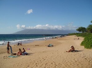 Uma praia com pessoas tomando sol e nadando, uma montanha ao fundo e um céu claro.