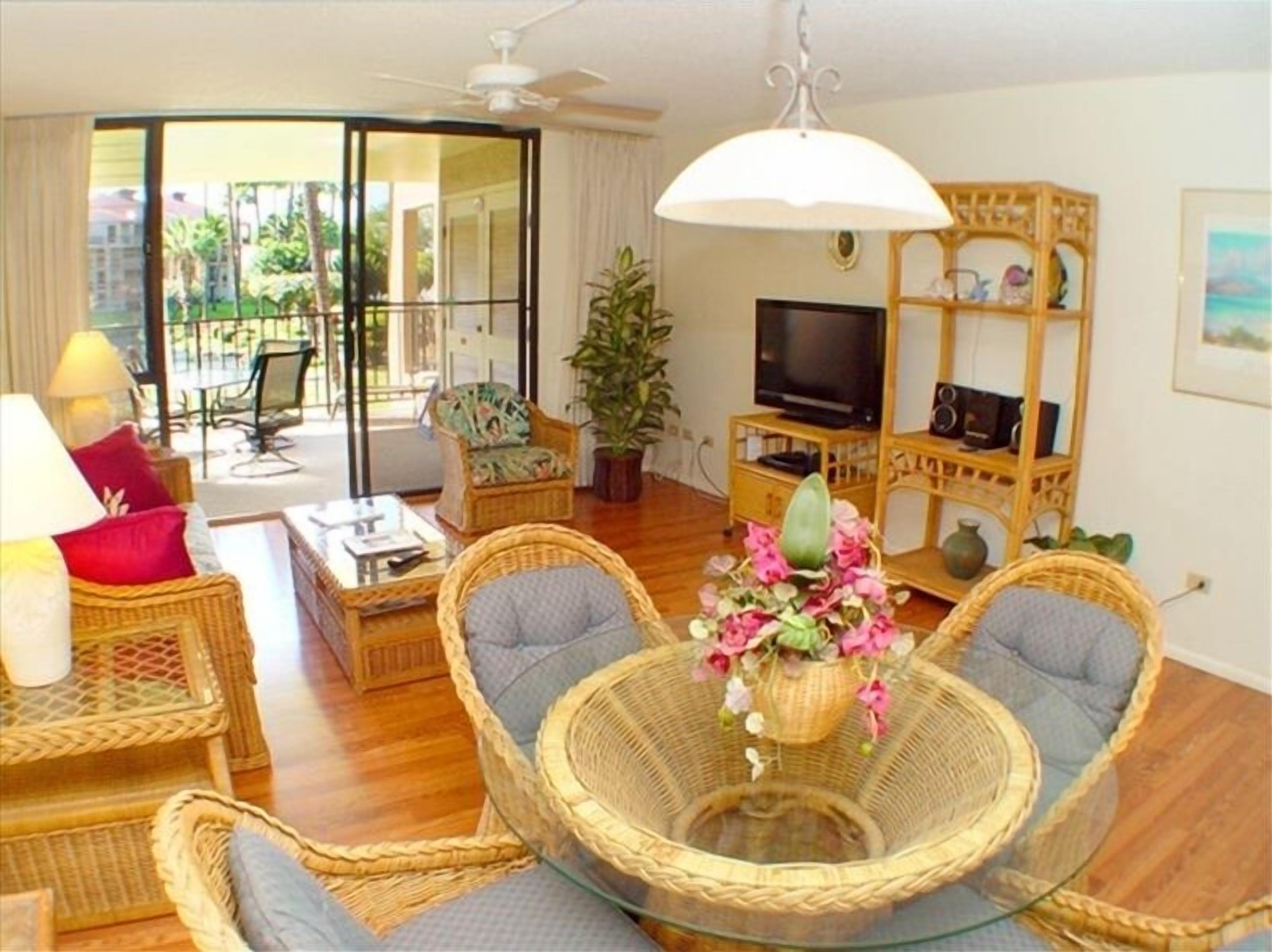 A living room with wicker furniture, a glass coffee table, a TV, and a balcony with a view of palm trees.