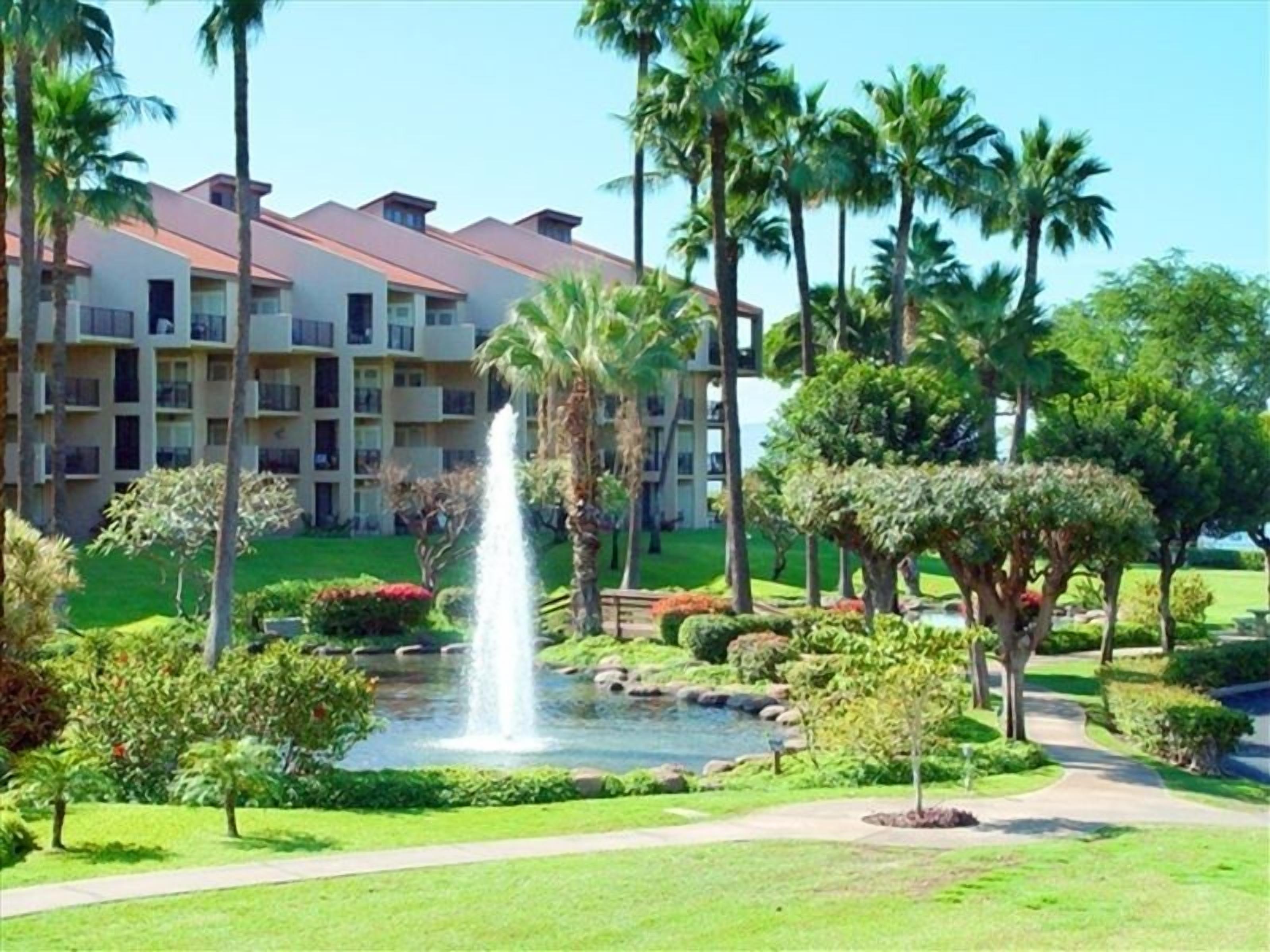 A hotel with a central fountain, surrounded by well-maintained lawns and palm trees.