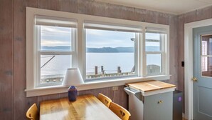 Ein Essbereich mit Holztisch und Stühlen, einer Lampe und Blick auf ein Gewässer durch große Fenster.