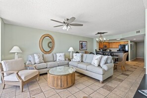 A living room with a sectional sofa, two armchairs, a coffee table, a ceiling fan, a chandelier, a dining area, and a kitchen in the background.