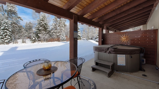 Un patio abrité avec une table et des chaises en verre, un jacuzzi et une vue sur un paysage enneigé.