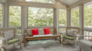 A covered porch with wicker furniture, a bench with cushions, and a view of trees through large windows. - Beautiful 4BR Rehoboth Beach Home w/ Gorgeous Porch (Rehoboth Beach)