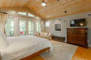Une chambre spacieuse avec un grand lit, un ventilateur de plafond en bois, un téléviseur fixé au mur et un coin salon confortable avec un canapé et des fauteuils.