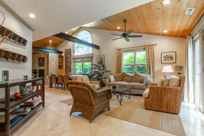 A spacious living room with a high ceiling, large windows, and a variety of seating options including a sofa, chairs, and a wicker armchair.