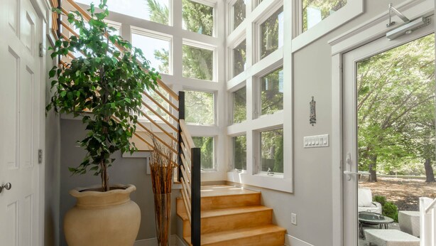 A staircase with a wooden handrail, a large potted plant, and a view of outdoor seating through a glass door. - Beautiful 4BR Rehoboth Beach Home w/ Gorgeous Porch (Rehoboth Beach)