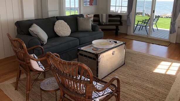 Un salon avec un canapé gris foncé, deux fauteuils en bois, une petite table avec un plateau, et une grande fenêtre donnant sur la mer.