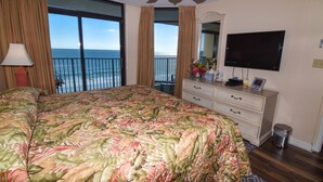 A bedroom with a large bed, a dresser, a TV, and a balcony with a view of the ocean.