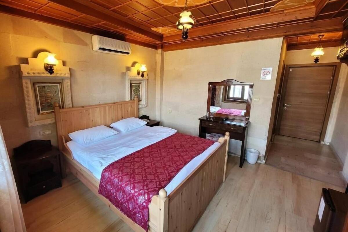 Un lit en bois recouvert d’une couverture rouge, une table de chevet avec une lampe, un miroir et une porte, dans une pièce dont le sol et le plafond sont en bois.
