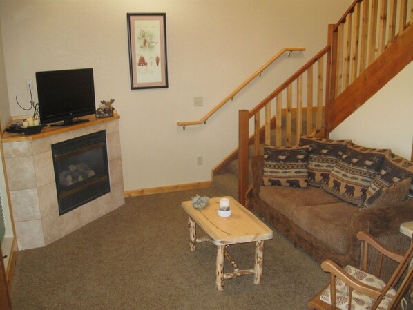 A living room with a fireplace, a television on a mantel, a sofa with patterned cushions, and a wooden staircase.