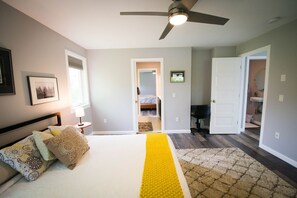 Une chambre à coucher avec un lit, un ventilateur de plafond, une porte donnant sur une salle de bain et un couloir.